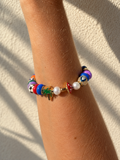 A colorful beaded millie madness bracelet with evil eye charms, white pearls, and a green palm tree charm, shown worn on a person's wrist against a textured beige wall with soft shadows.