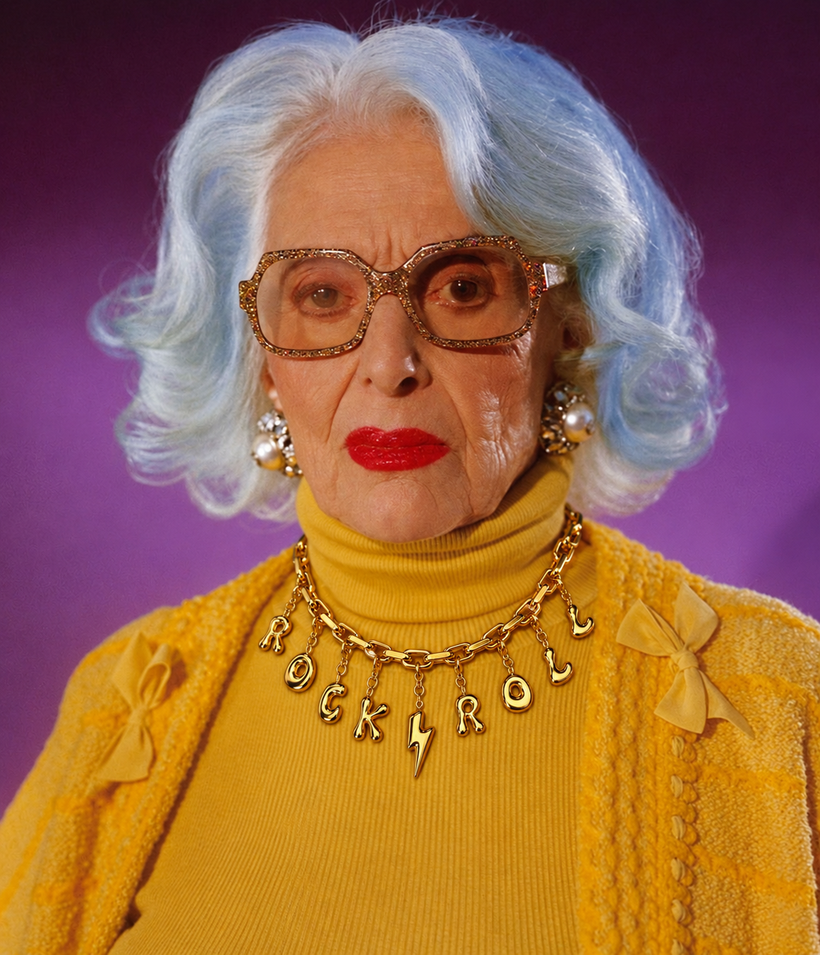 A woman with short, wavy light blue hair, wearing a bright yellow turtleneck sweater, matching yellow cardigan, and a gold millie madness necklace spelling 'ROCK ROLL' with a lightning bolt charm, set against a purple studio background.