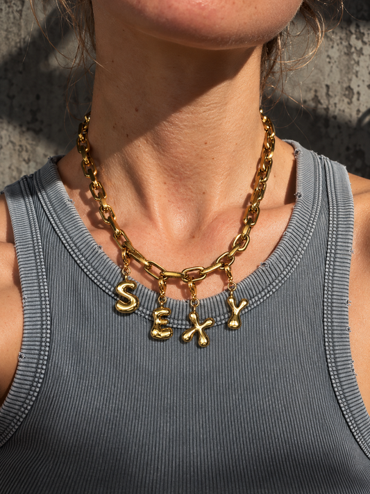 A close-up of a model wearing a chunky gold millie madness chain necklace with dangling letters spelling 'SEXY' in a ribbed gray tank top.