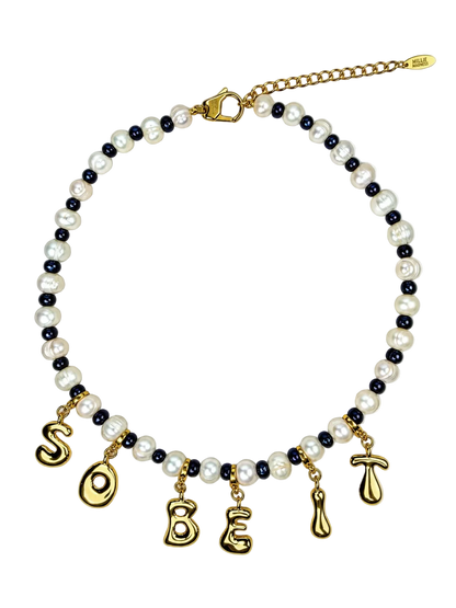 A beaded MILLIE MADNESS necklace featuring white faux pearls, dark navy blue beads, and gold-toned metal charms spelling 'SO BE' with additional gold charms, shown on a plain white background.