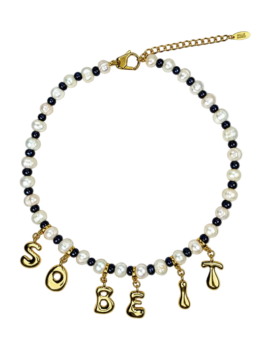 A beaded MILLIE MADNESS necklace featuring white faux pearls, dark navy blue beads, and gold-toned metal charms spelling 'SO BE' with additional gold charms, shown on a plain white background.