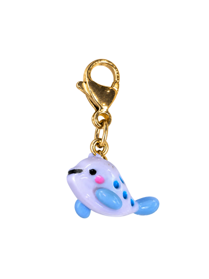 A small whale-shaped charm with a gold lobster clasp, featuring a pale lavender body, light blue flippers and spots, and pink and black facial details, isolated on a white background.