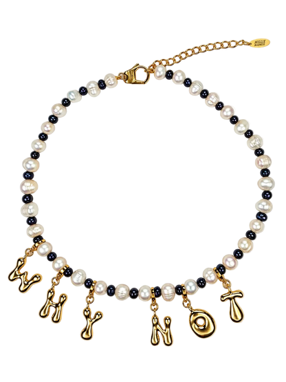A MILLIE MADNESS necklace featuring white baroque pearls and small black pearls, with dangling gold charms shaped like letters and symbols, shown on a plain white background