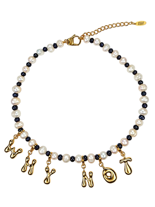 A MILLIE MADNESS necklace featuring white baroque pearls and small black pearls, with dangling gold charms shaped like letters and symbols, shown on a plain white background