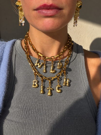 A close-up of a model wearing a layered millie madness gold chain necklace with dangling letter charms spelling 'WILD BITCH' in gold, along with matching gold drop earrings.