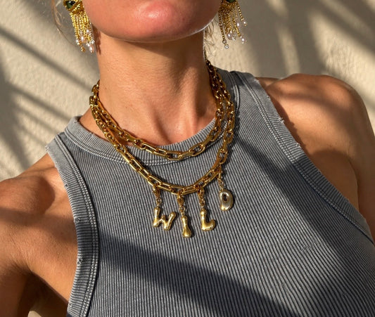 A close-up of a person wearing a layered millie madness gold necklace with dangling letter charms spelling 'WILD' and matching gold drop earrings, paired with a gray ribbed tank top against a sunlit wall.
